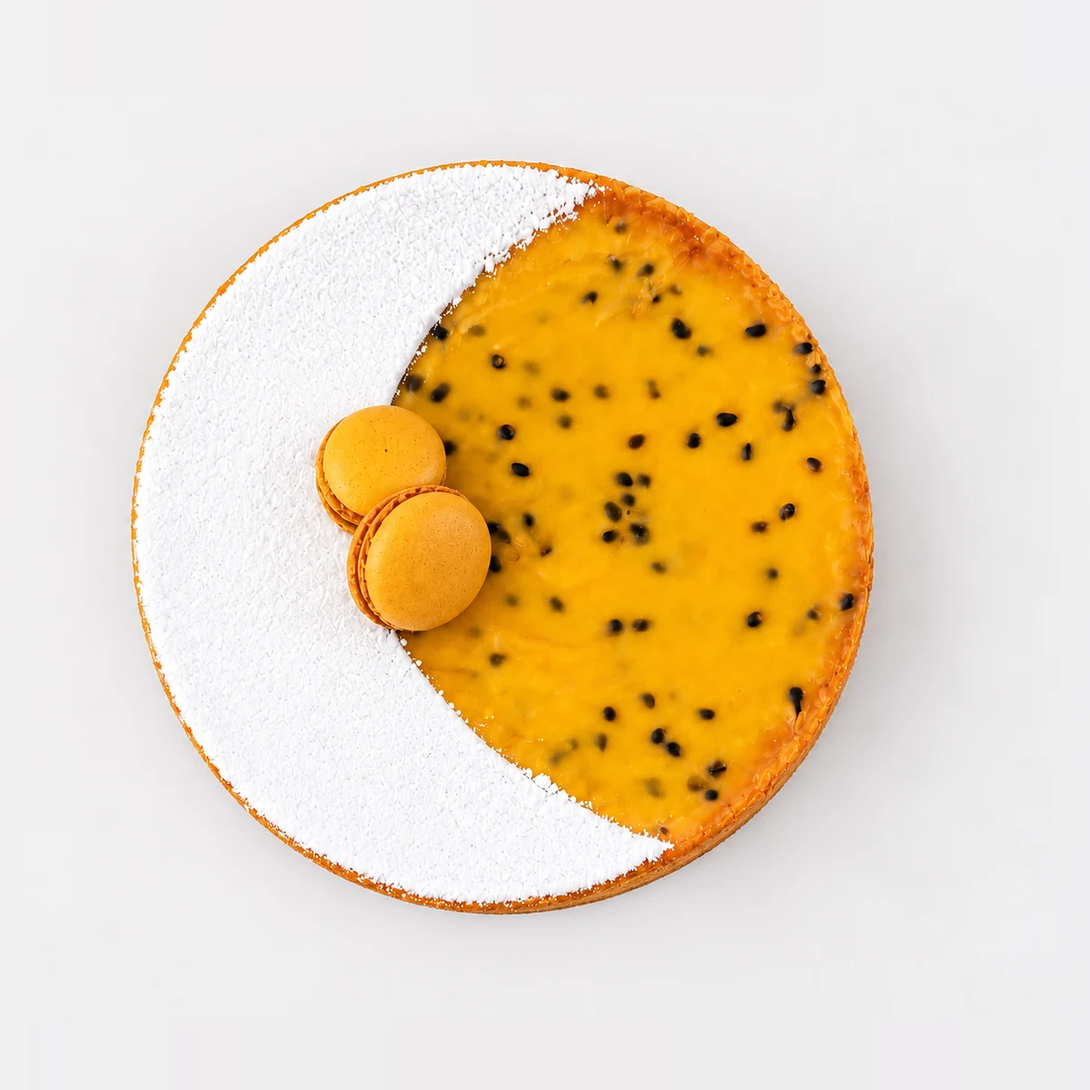 Passionfruit tart from above: a crescent of icing sugar beside glossy passionfruit curd with seeds and two macarons.