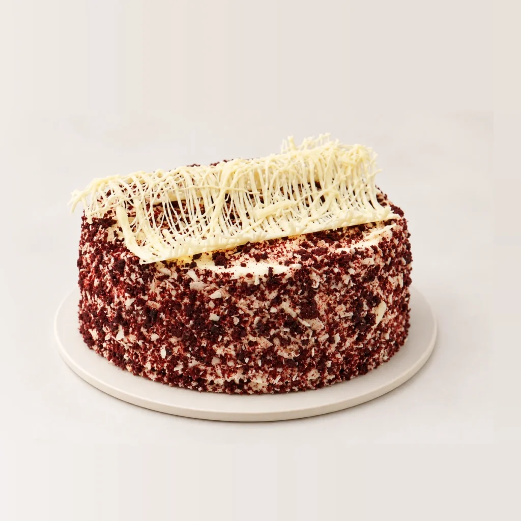 Red velvet cake coated in red velvet crumb, topped with a white chocolate decoration.