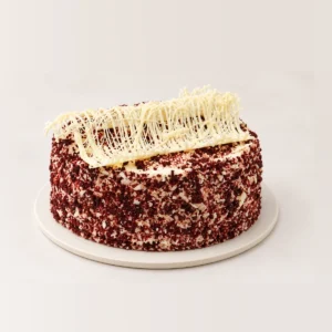 Red velvet cake coated in red velvet crumb, topped with a white chocolate decoration.