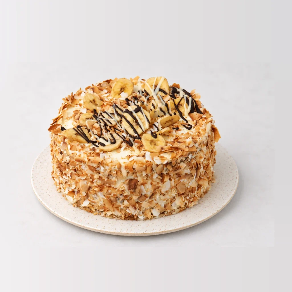 Tropical banana and coconut cake coated in toasted coconut flakes, topped with banana slices and a chocolate drizzle.
