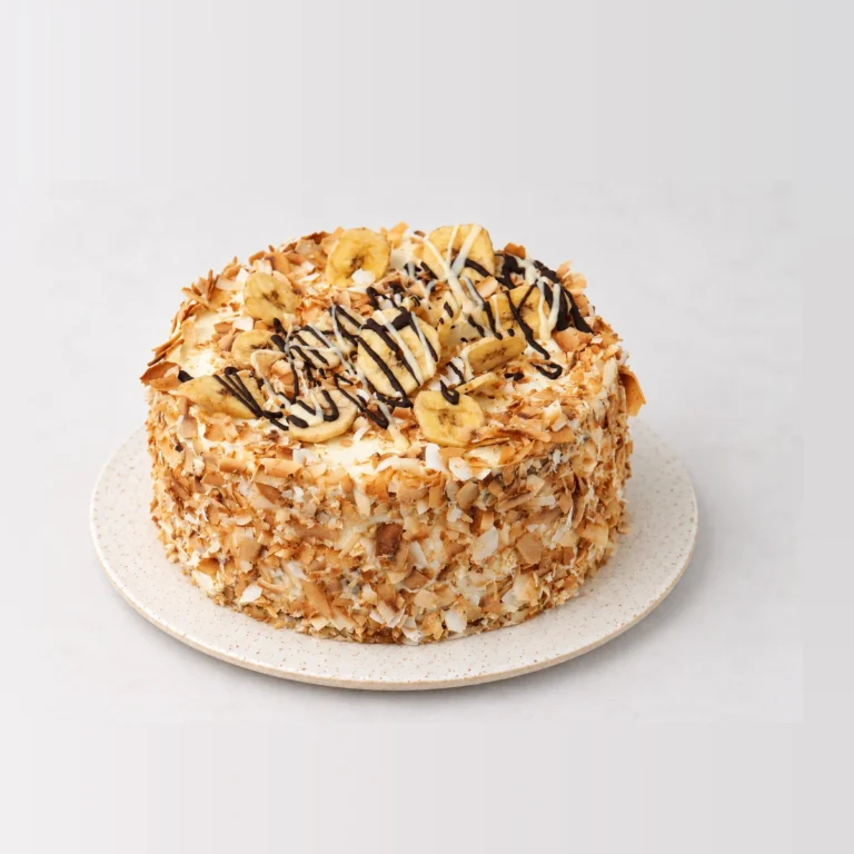 Tropical banana and coconut cake coated in toasted coconut flakes, topped with banana slices and a chocolate drizzle.