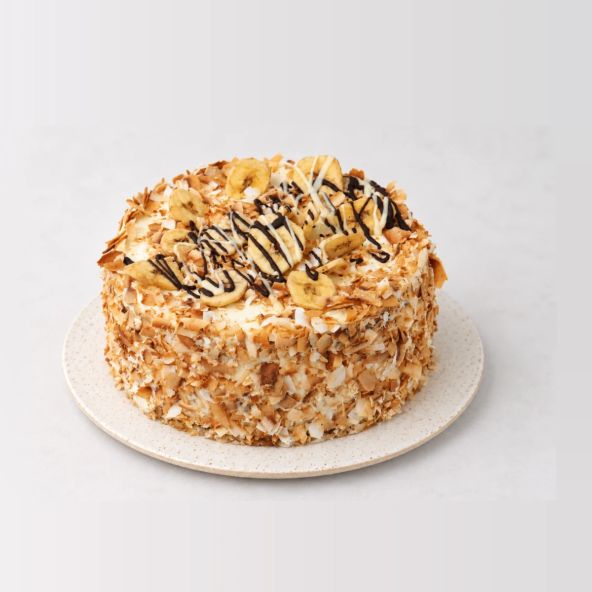 Tropical banana and coconut cake coated in toasted coconut flakes, topped with banana slices and a chocolate drizzle.